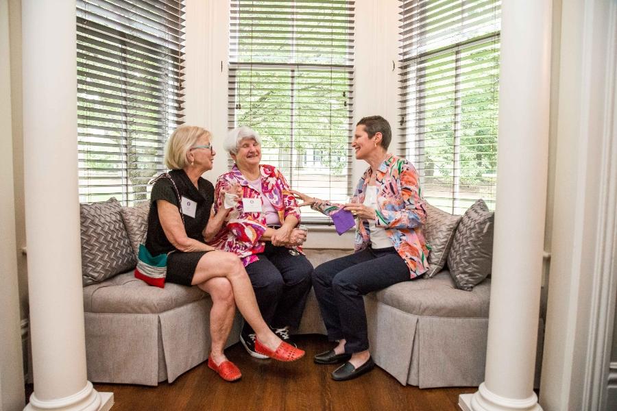 Three people talk while holding drinks and sitting on a sectional couch inset of a window well. 